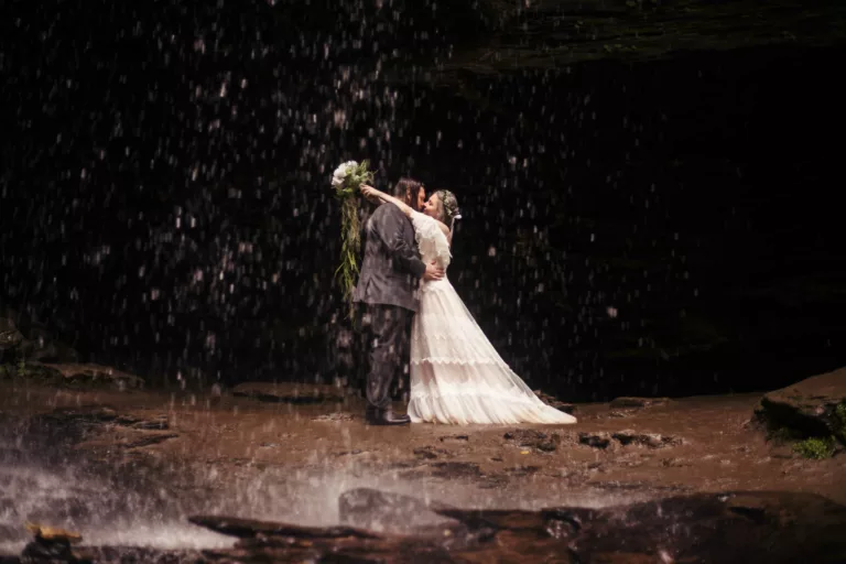 Bride and Groom standing in a light drizzle of a waterfall. He is holding her waist and she is hugging his neck.